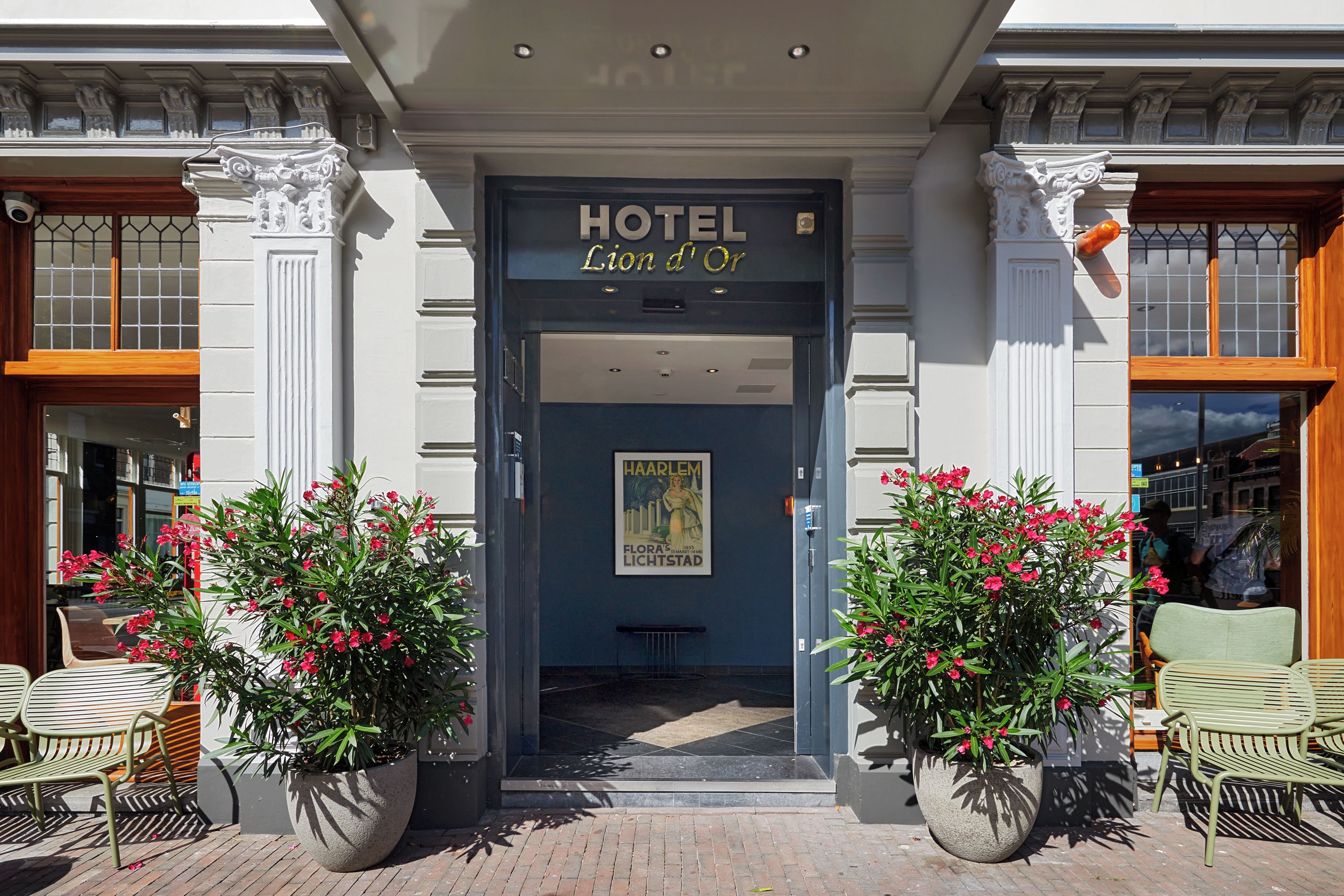 Ein Hotelzugang mit einer dunkelblauen Tür, einem Schild mit der Aufschrift 'HOTEL Lion d'Or' und je einer großen Pflanze mit roten Blüten auf jeder Seite.
