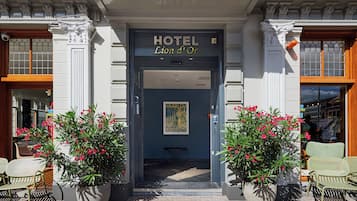 Ein Hotelzugang mit einer dunkelblauen Tür, einem Schild mit der Aufschrift 'HOTEL Lion d'Or' und je einer großen Pflanze mit roten Blüten auf jeder Seite.