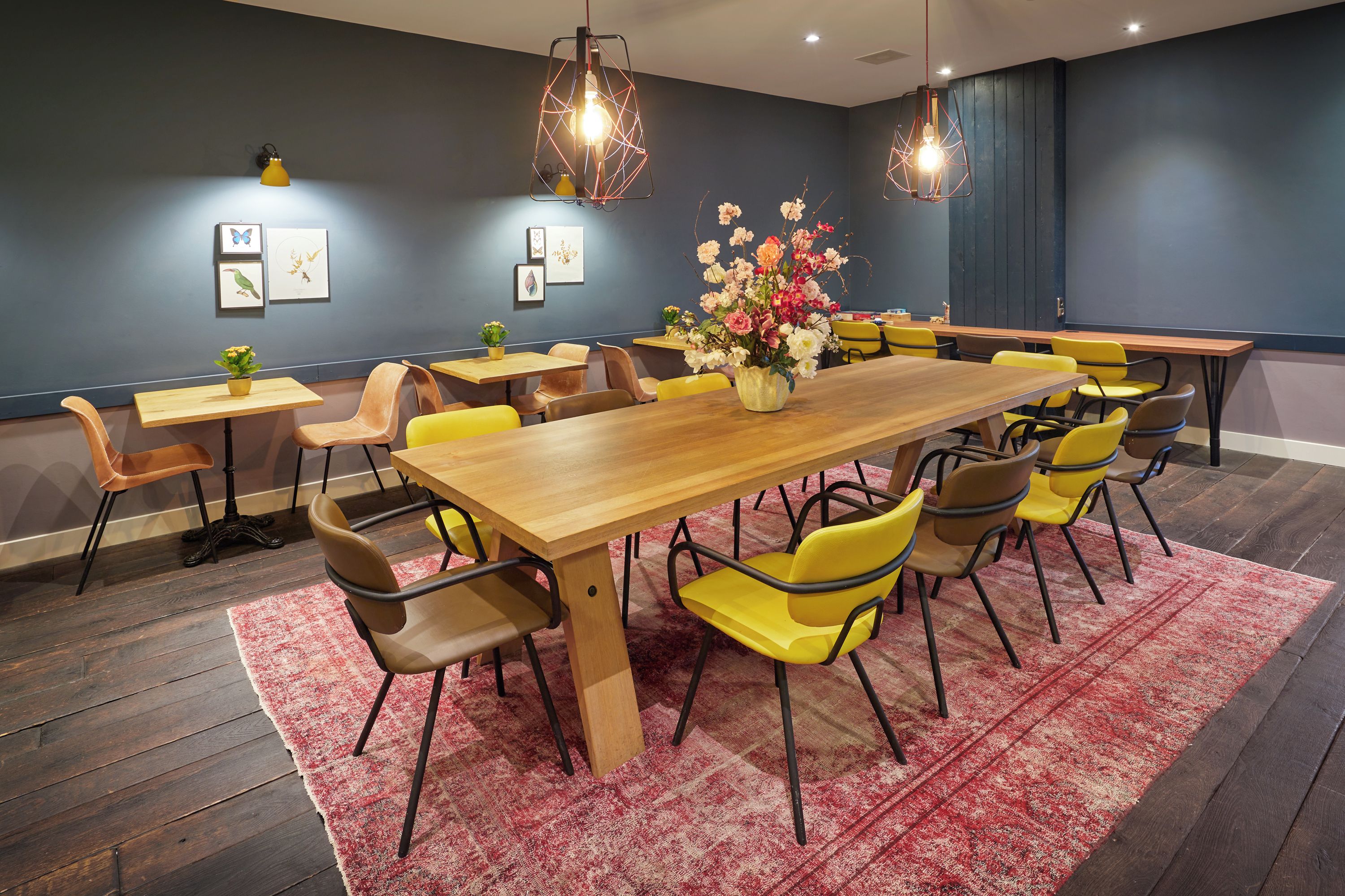 A conference room with a large wooden table surrounded by yellow chairs, a red and white patterned rug, and wall-mounted pendant lights.