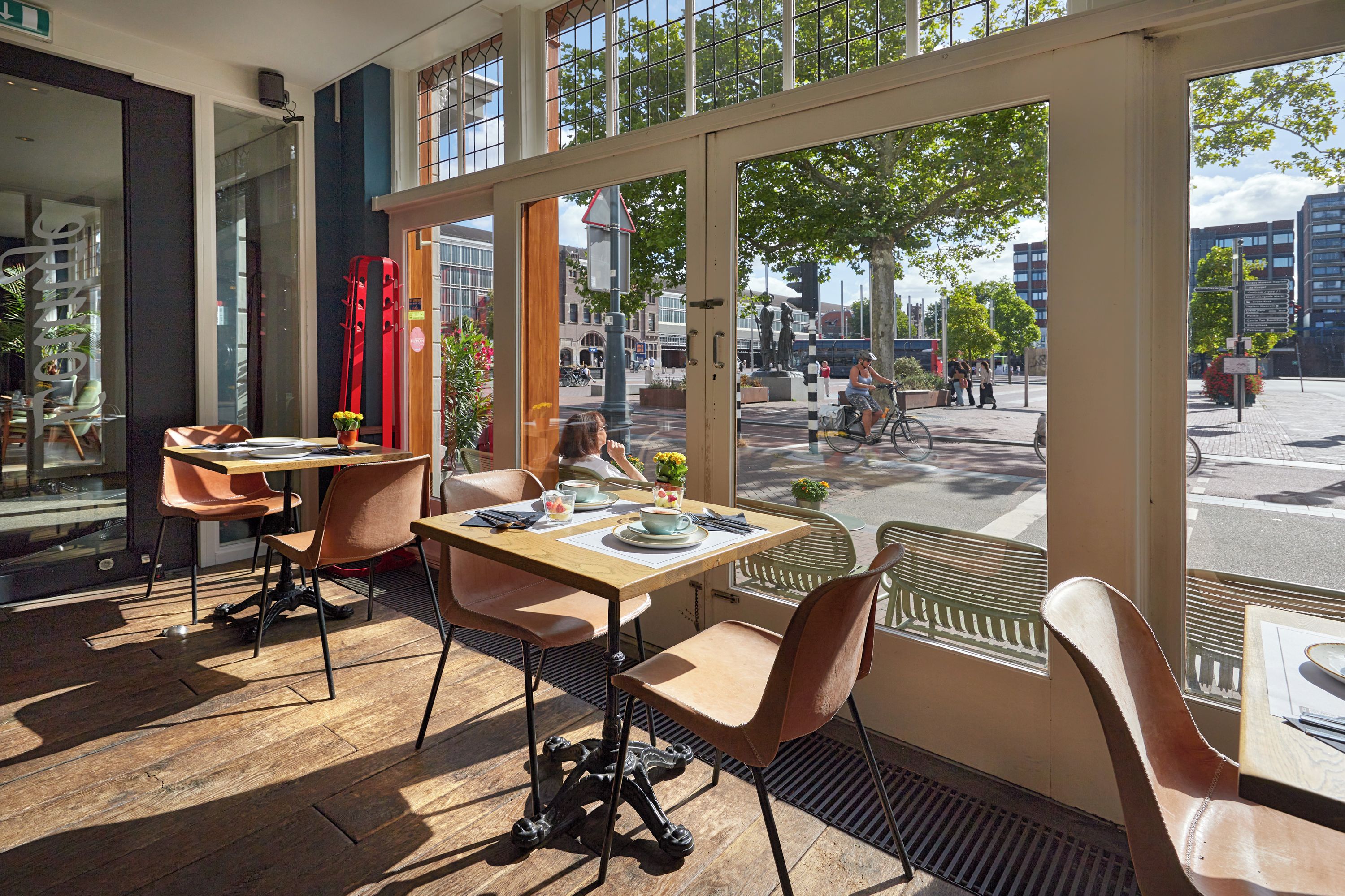 An interior with large windows, a dining area with tables and chairs, and a view of the street outside.