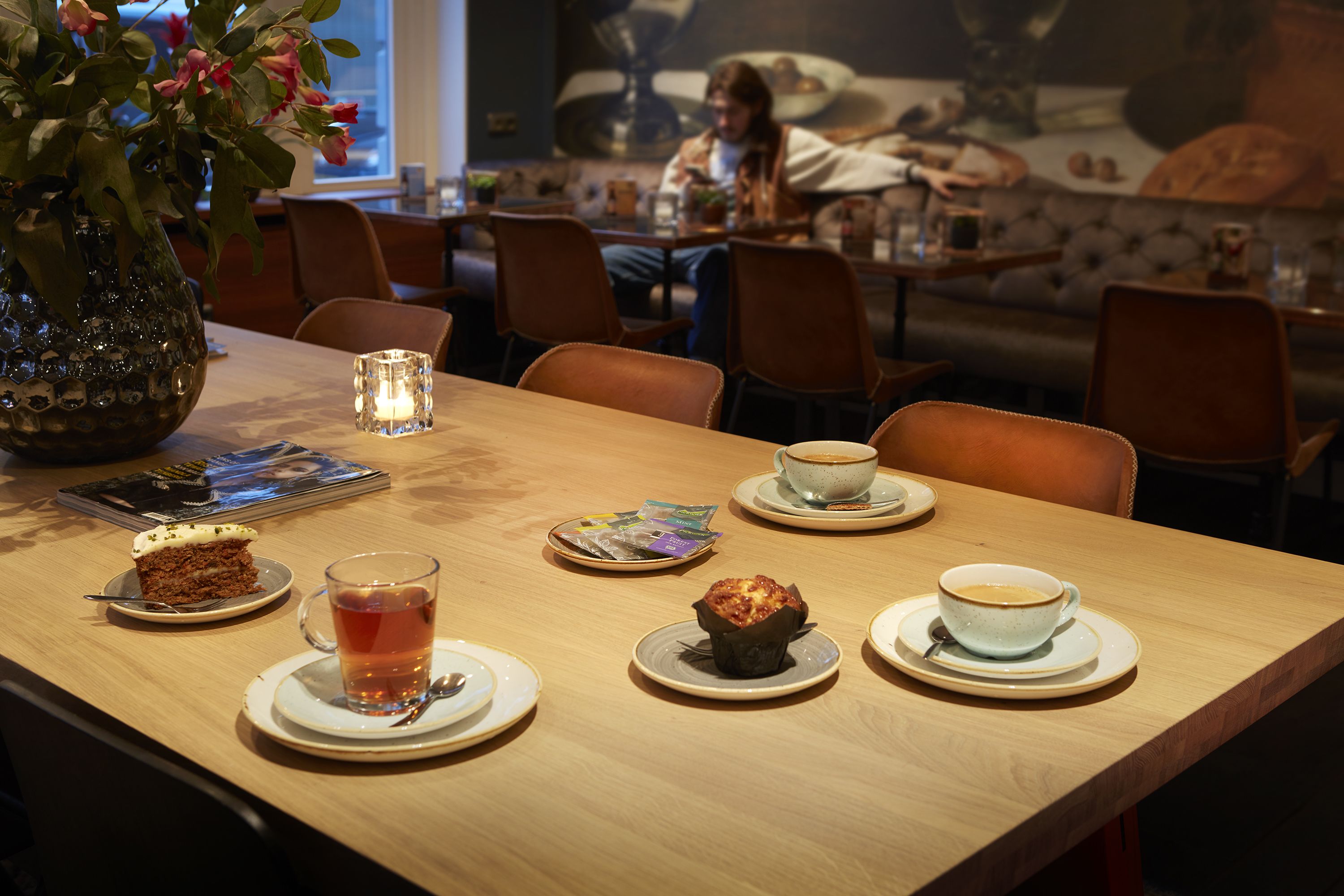 A wooden table with a vase of flowers, a candle, and various food items including a slice of cake, a muffin, and a cup of tea.