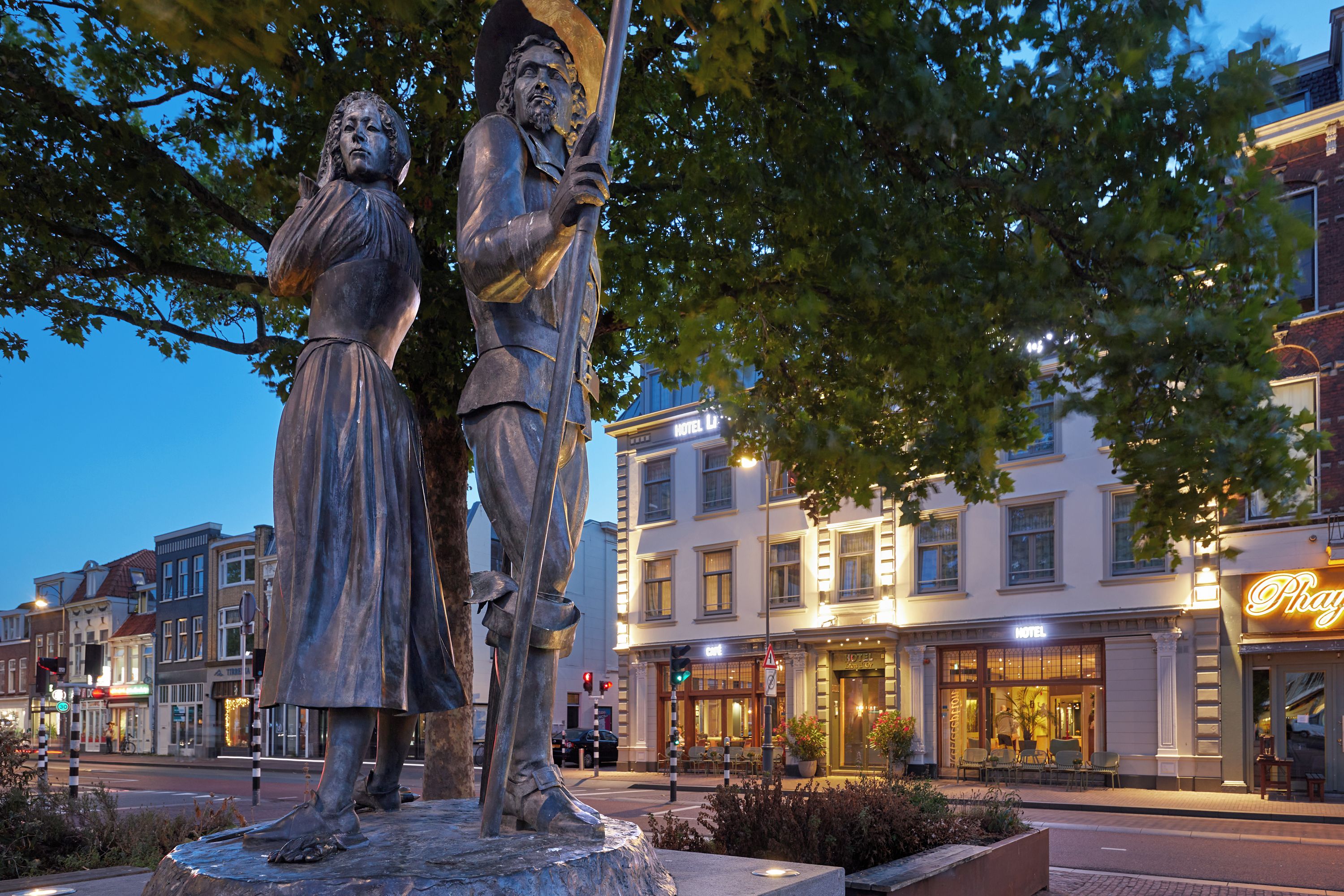 En staty som föreställer två figurer, placerad på ett offentligt torg med ett hotell och butiker i bakgrunden.