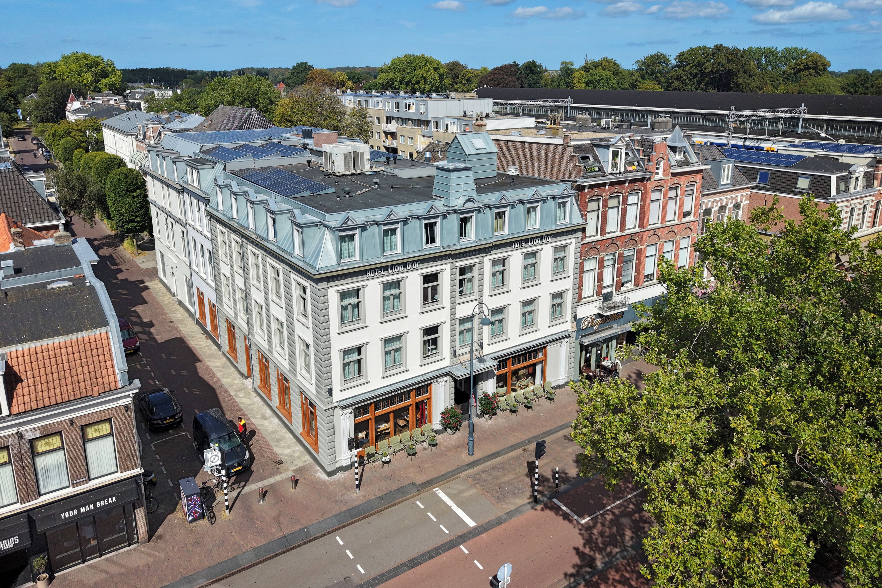 Un hotel de varios pisos con un estilo arquitectónico europeo tradicional, que presenta una entrada destacada en esquina y una terraza en la azotea con paneles solares.
