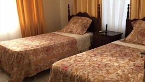 A room with two beds, each with a floral bedspread, a wooden headboard, and a nightstand with a lamp. The room has a tiled floor and a window with curtains. - Ocho Rios Golden View (Kingston)