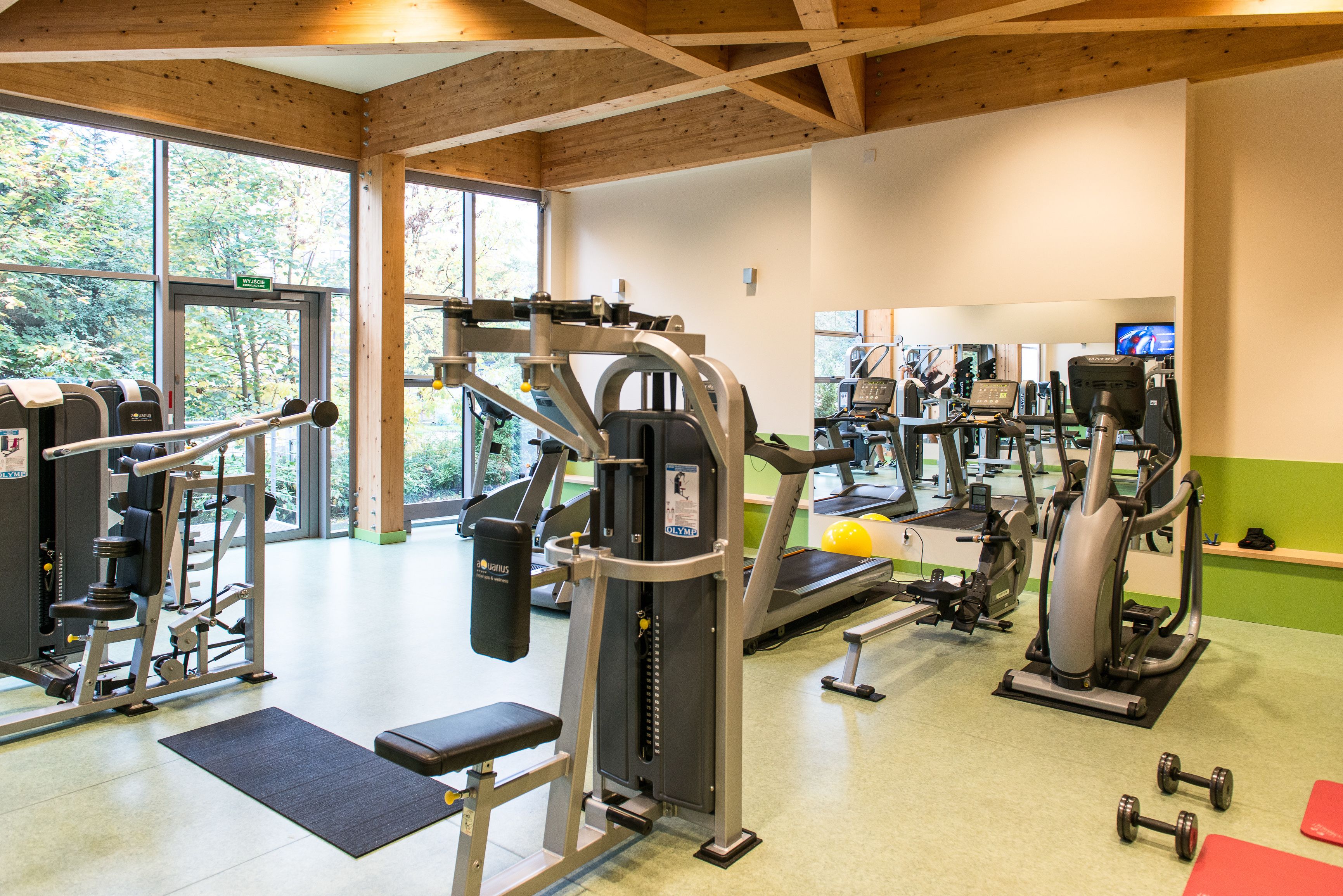 Ein Fitnessstudio mit verschiedenen Trainingsgeräten, großen Fenstern und einer grünen Wand.