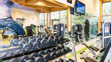 A gym with exercise equipment, including dumbbells and weight machines, and large windows with a view of trees.