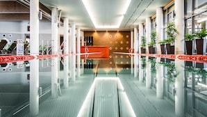 Un área de piscina interior moderna con suelo de vidrio transparente, pilares blancos y un mostrador de recepción al fondo.