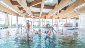 Una piscina interior con vigas de madera, agua cristalina y sillas verdes a los lados.