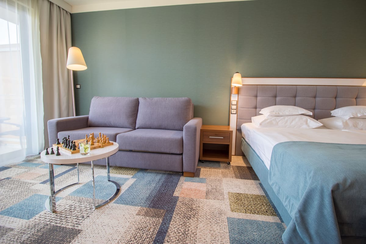 A hotel room with a sofa, a small table with a chess set, a bed with a blue bedspread, and a nightstand with a lamp.