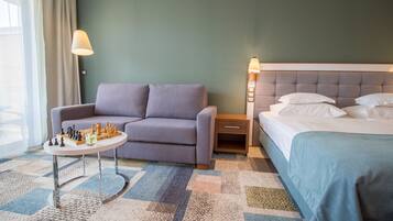 A hotel room with a sofa, a small table with a chess set, a bed with a blue bedspread, and a nightstand with a lamp.