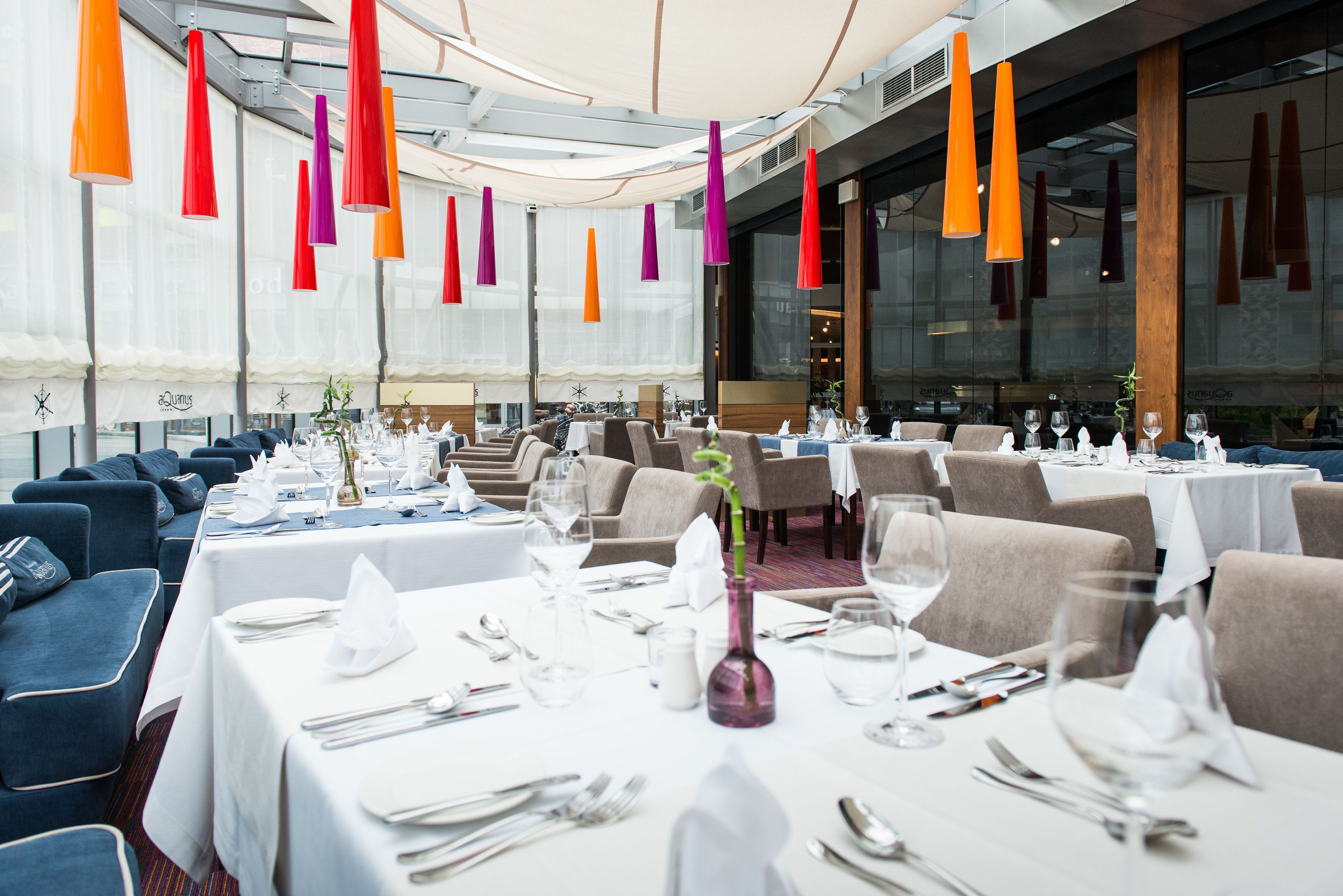 A restaurant with tables set for dining, featuring a modern interior with hanging pendant lights and a view of the cityscape.