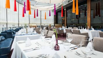 A restaurant with tables set for dining, featuring a modern interior with hanging pendant lights and a view of the cityscape.