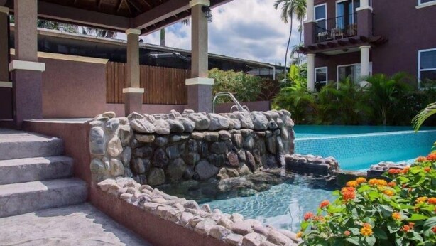 A stone hot tub with steps leading up to it, surrounded by a pool and a building with balconies.