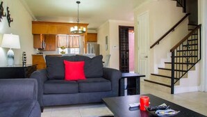 A living room with a sofa, a coffee table, and a TV. There is a kitchen in the background with wooden cabinets and a chandelier. A staircase is visible to the right.