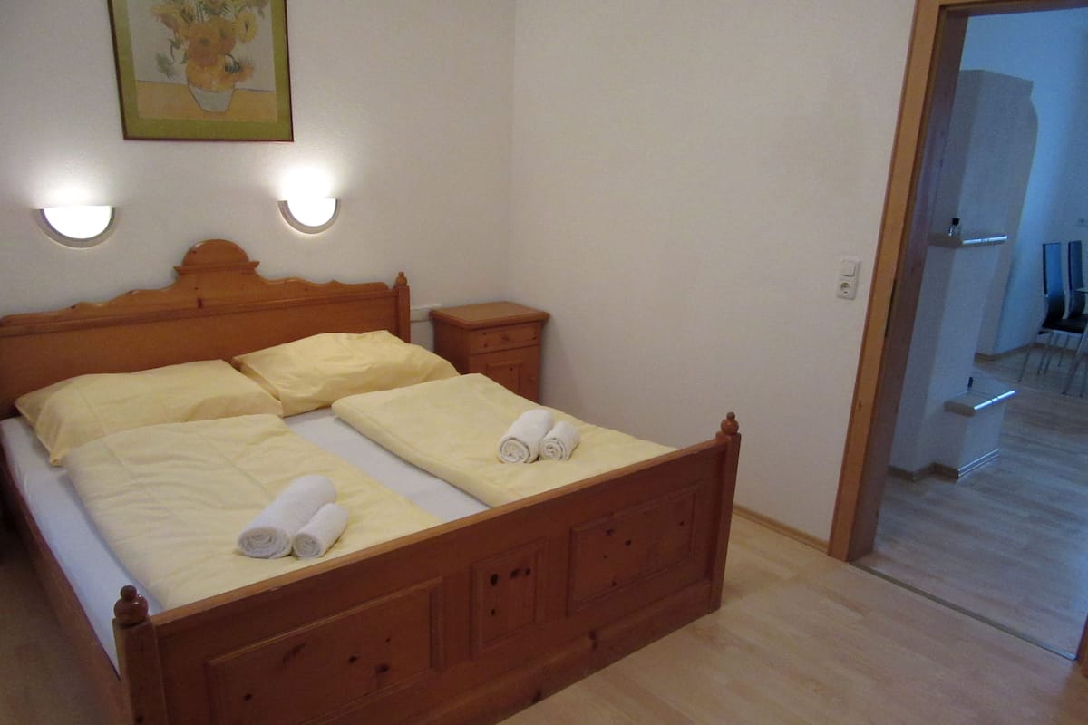 Uma cama de madeira com duas almofadas e um cobertor, uma mesinha de cabeceira e um aplique na parede.