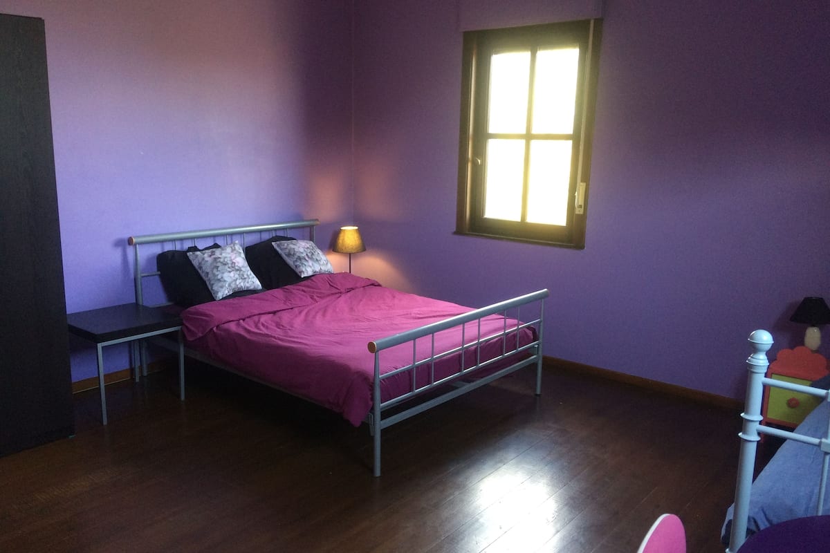 A bedroom with a metal bed frame, a bedside table, a lamp, and a window with a view of the outside.