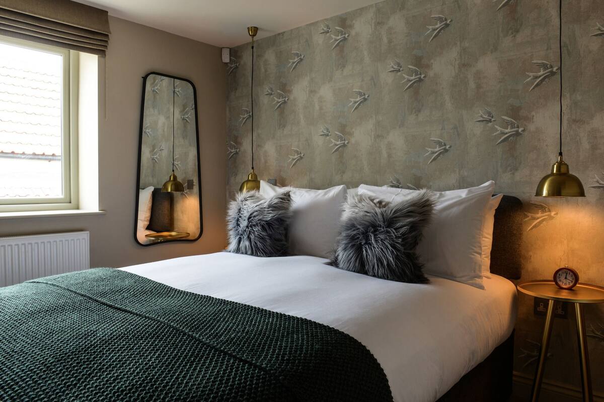 A neatly made bed with a green blanket, a large mirror, and a wall with bird designs.