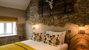 Un dormitorio con pared de piedra, cama con almohadas estampadas, manta amarilla y dos lámparas de pared.