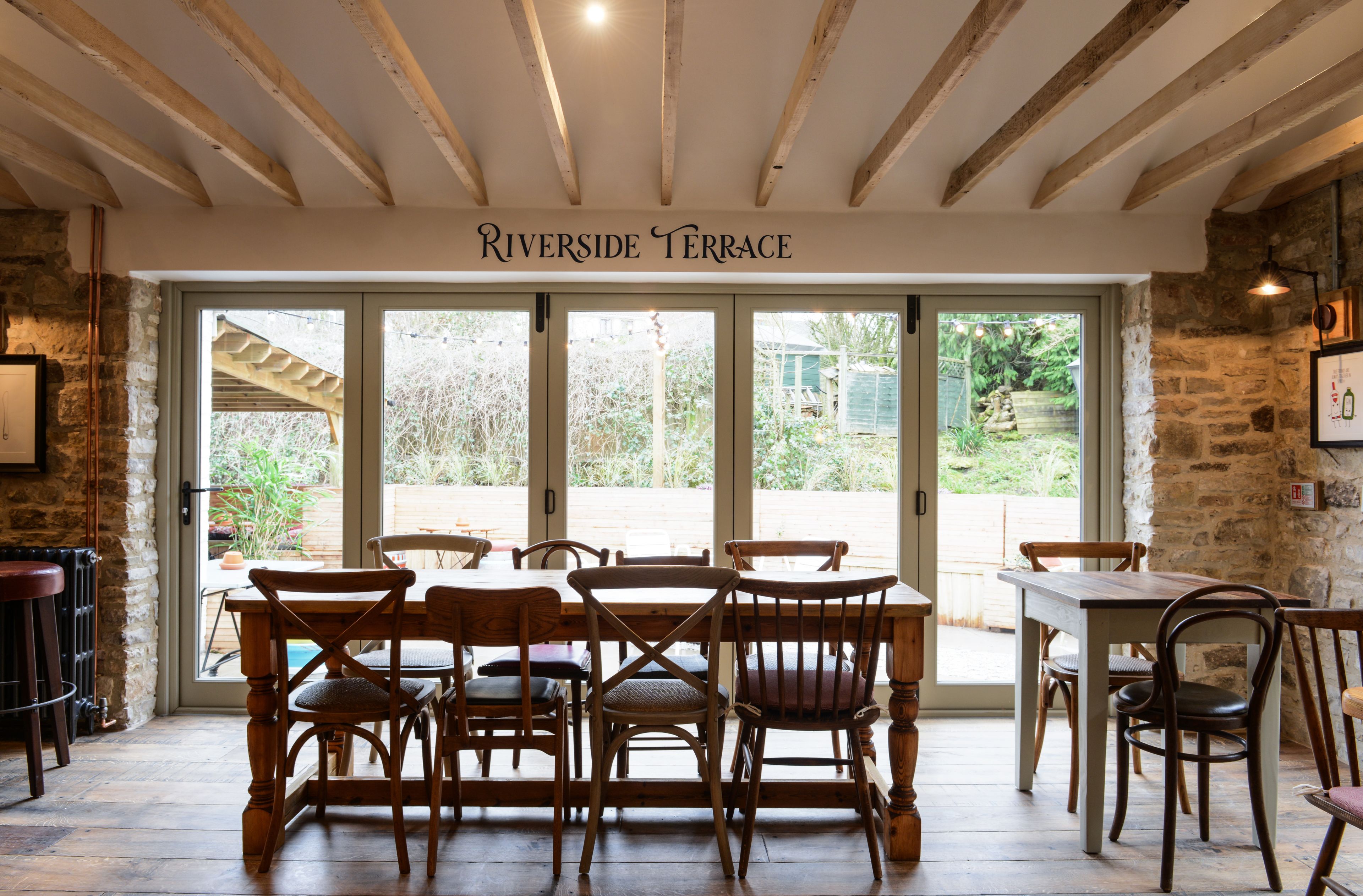 A dining area with wooden tables and chairs, a stone wall, and a sign reading "Riverside Terrace".