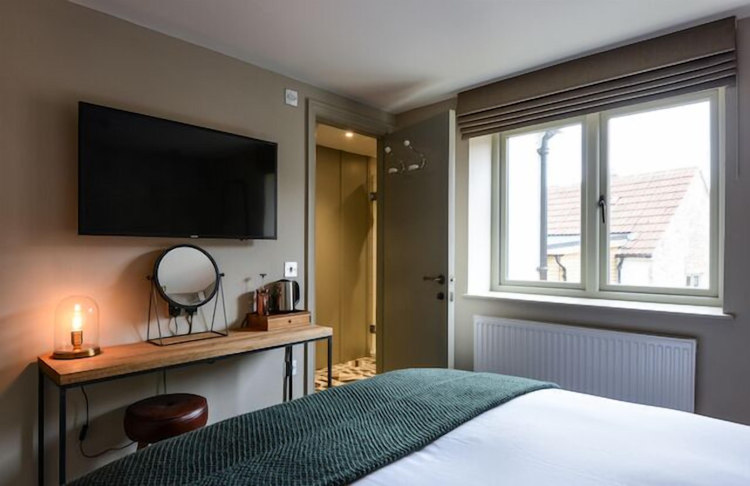 A bedroom with a bed, a desk with a lamp, a mirror, and a TV mounted on the wall.