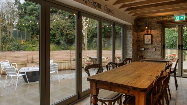 Una mesa de comedor de madera con sillas, ventanas amplias y una pared de piedra con un letrero que dice "Riverside Terrace".