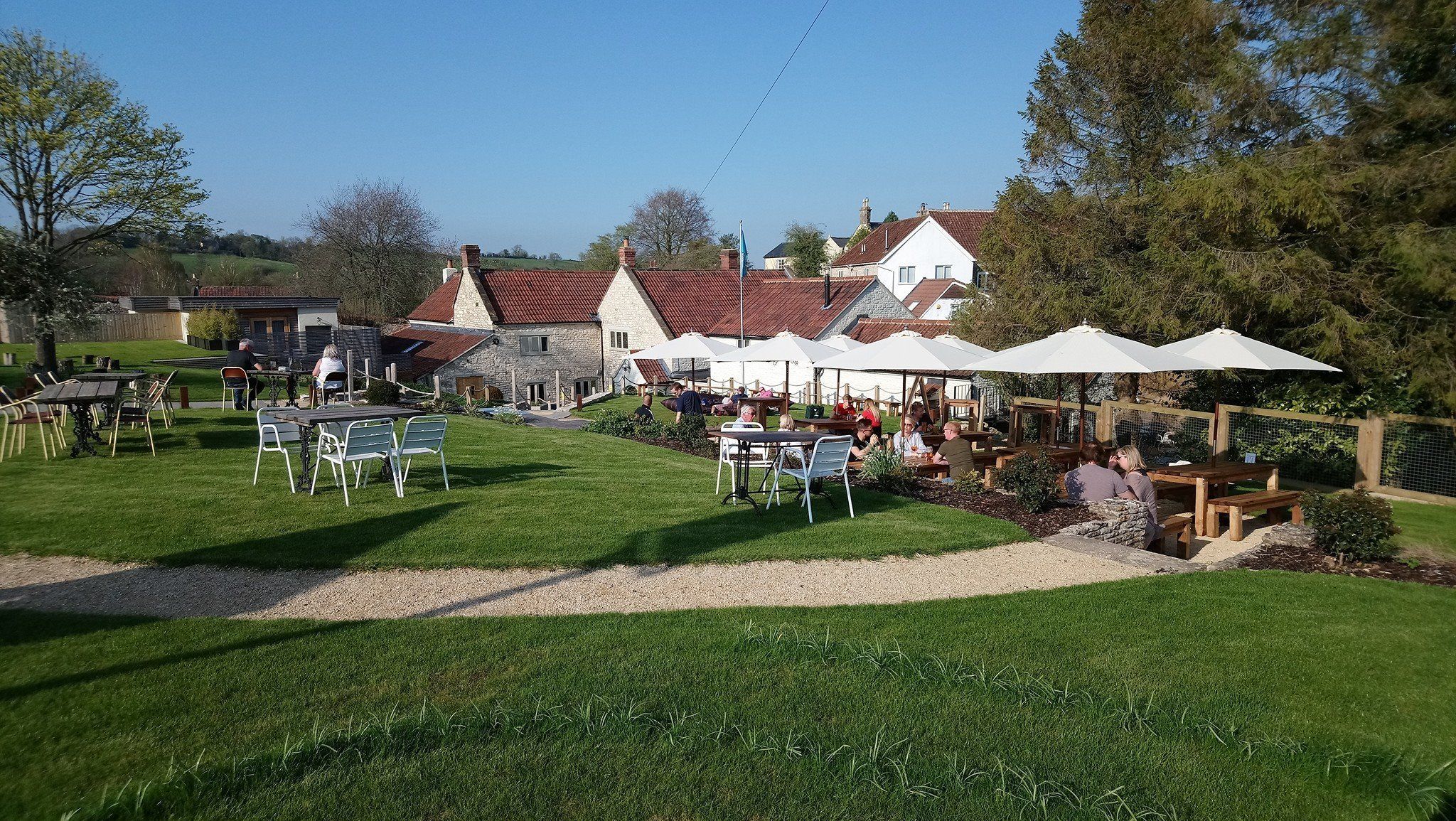 An outdoor dining area with tables and chairs, surrounded by a grassy lawn and a stone building with a red roof.
