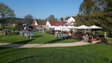 An outdoor dining area with tables and chairs, surrounded by a grassy lawn and a stone building with a red roof.