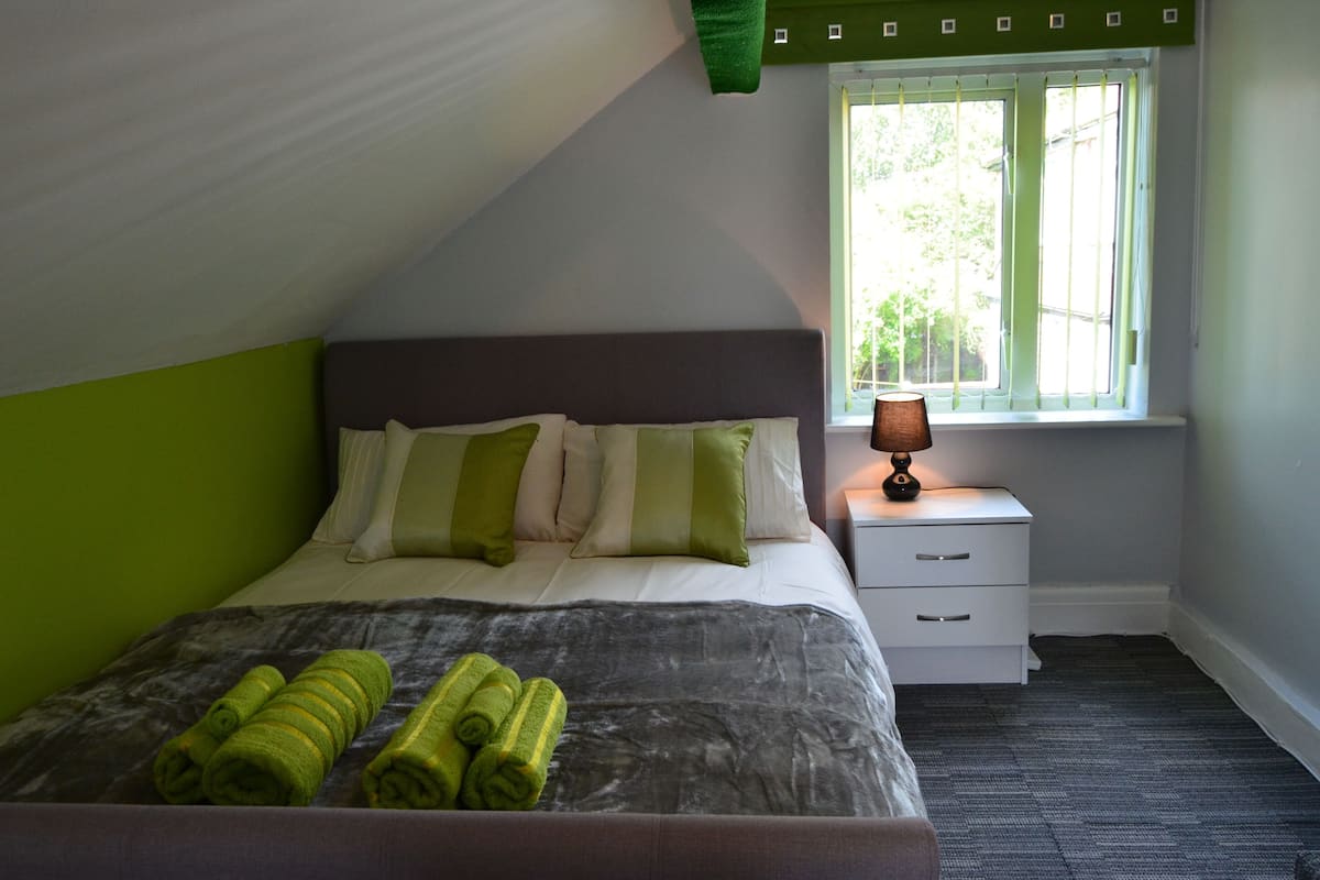 A bedroom with a bed, green pillows, a bedside table with a lamp, and a window with curtains.