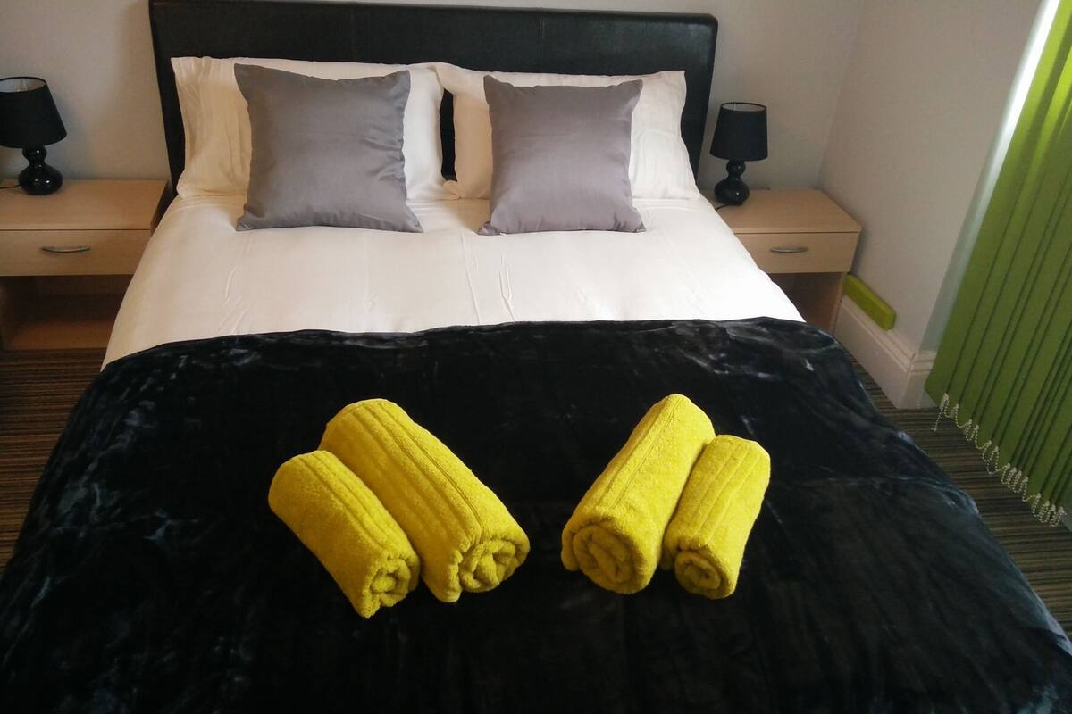 A hotel room with a bed, two bedside tables, a black velvet bedspread, and two rolled towels on the bed.