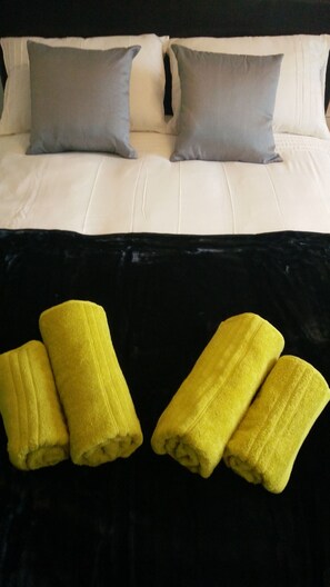 A bed with white linens and two gray pillows. Below the bed, there are two rolled-up yellow towels on a black surface. - Old Trafford Guest House (Manchester)