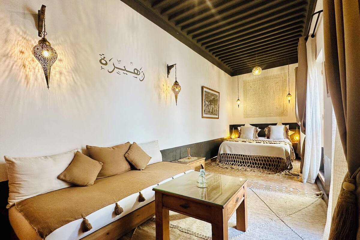 A room with a sofa, a wooden table, a bed, wall-mounted lights, and a decorative wall piece.
