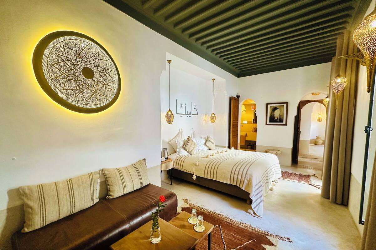 A bedroom with a bed, a sofa, a coffee table with a vase, and a decorative wall piece.