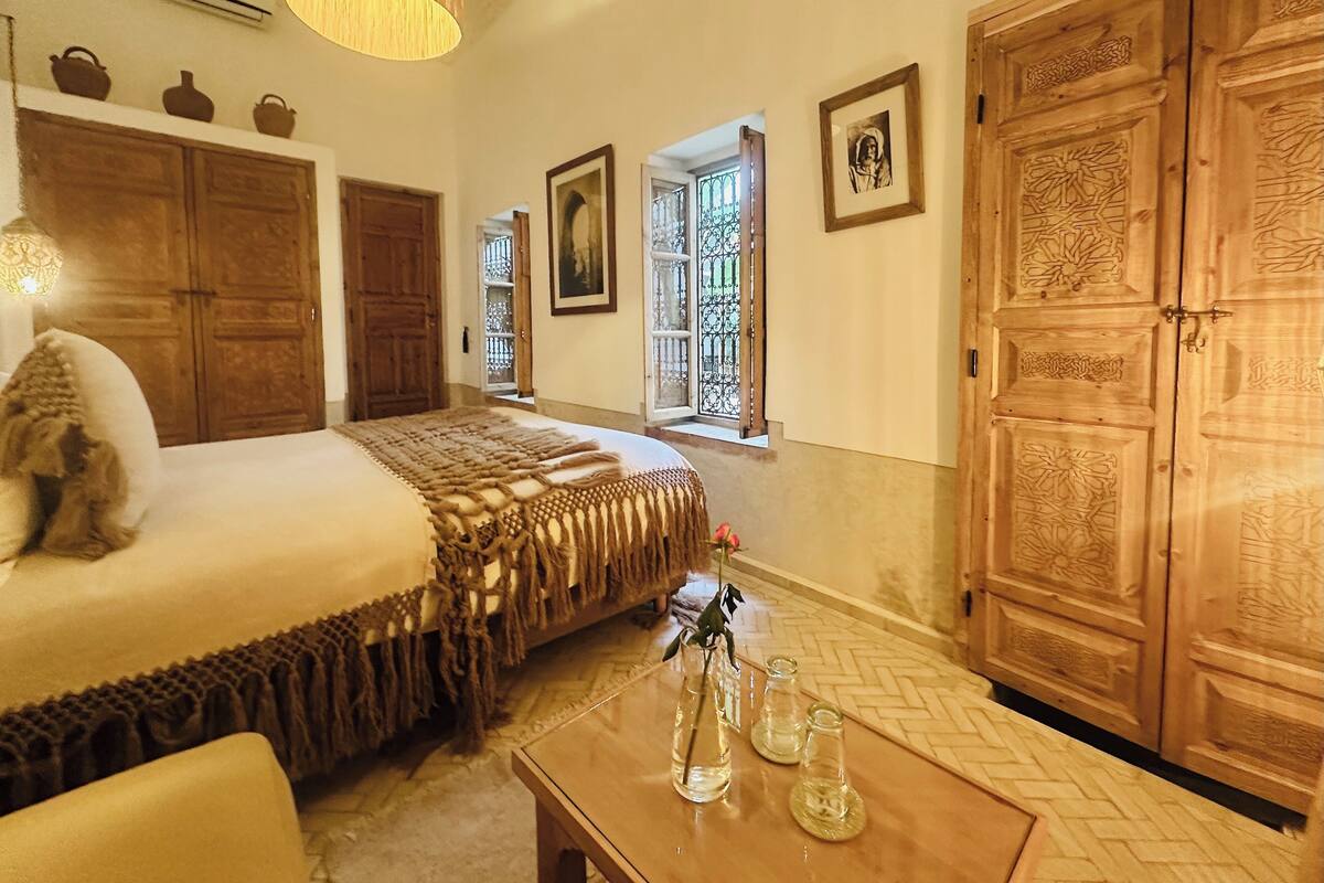 A bedroom with a large bed, wooden wardrobe, and a small table with decorative items.