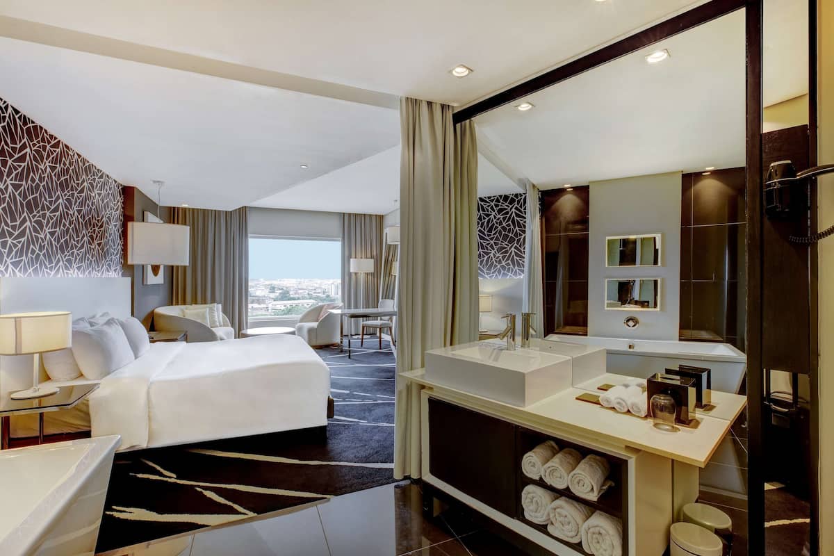 A hotel room with a large bed, a desk with a lamp, a vanity with a mirror, and a view of the city.