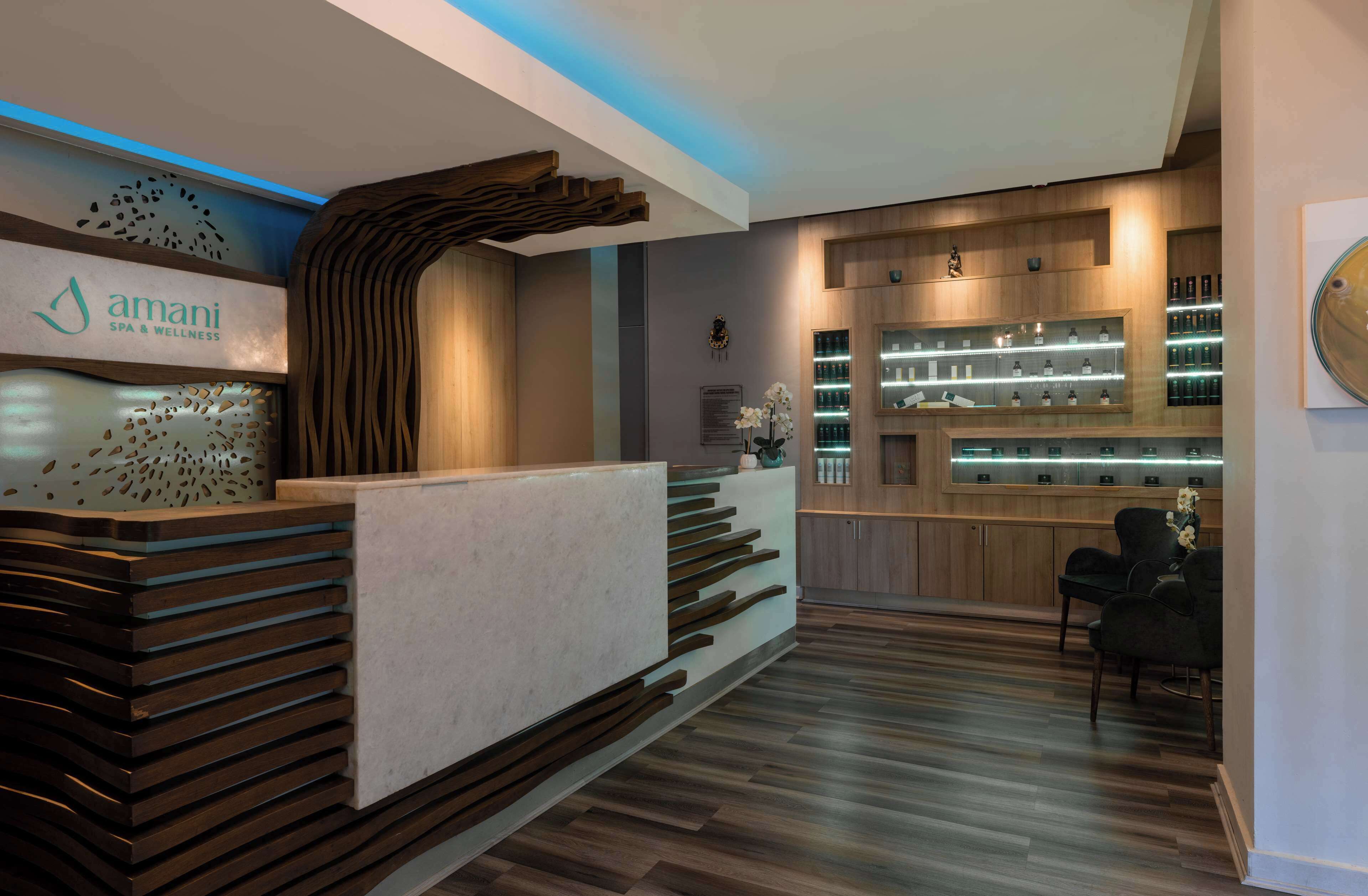 Un área de recepción moderna con un mostrador de madera, un panel mural con el logo de 'Amani Spa & Wellness' y una estantería con diversos productos.