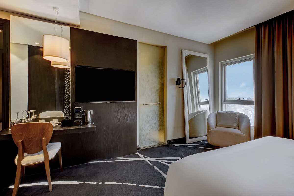 A hotel room with a bed, a desk with a flat-screen TV, a chair, and a large window.