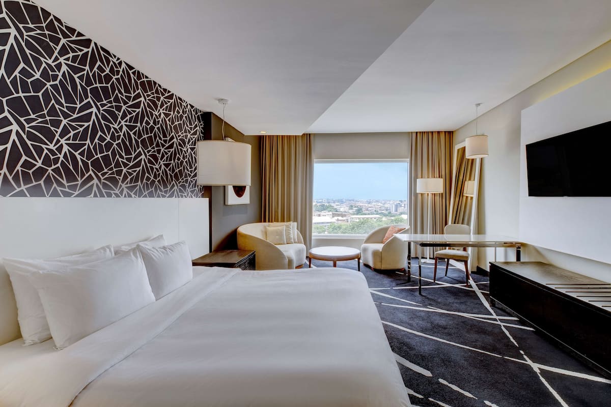 A hotel room with a large bed, a desk, two chairs, a TV, and a view of the city.