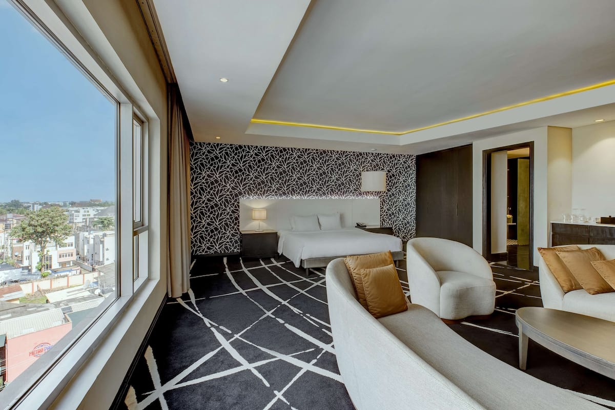 A hotel room with a large window, a bed with a white headboard, a sofa, and a coffee table.