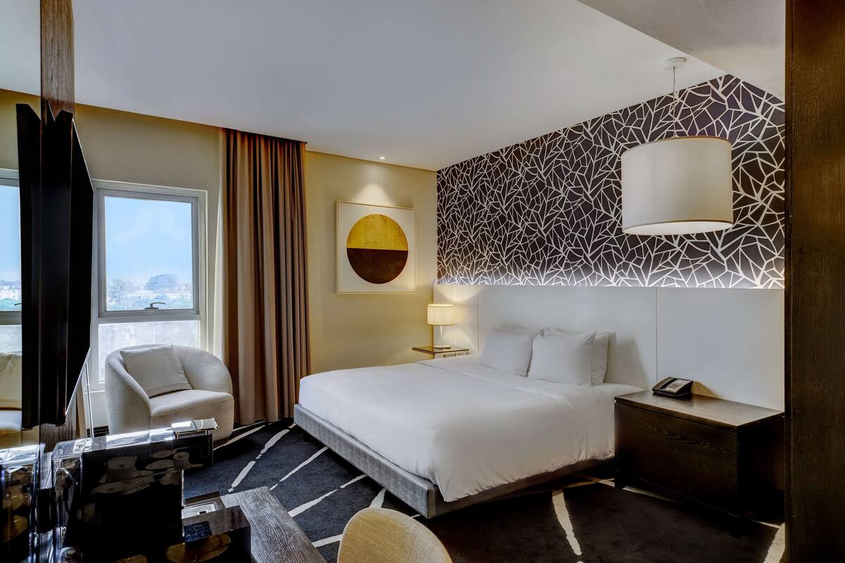 A hotel room with a large bed, a desk, a chair, a lamp, and a view of the city.