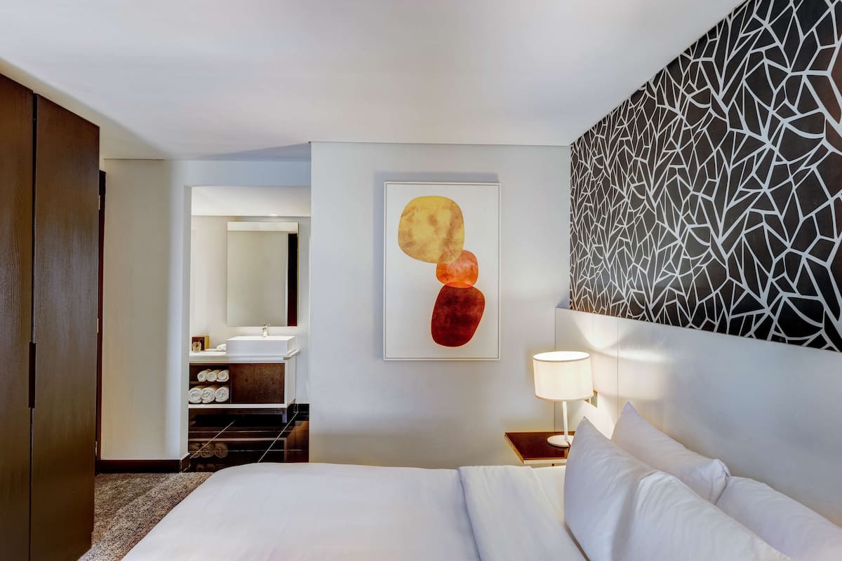 A hotel room with a bed, a nightstand with a lamp, a wall-mounted abstract art piece, a bathroom with a sink and mirror, and a patterned wall.