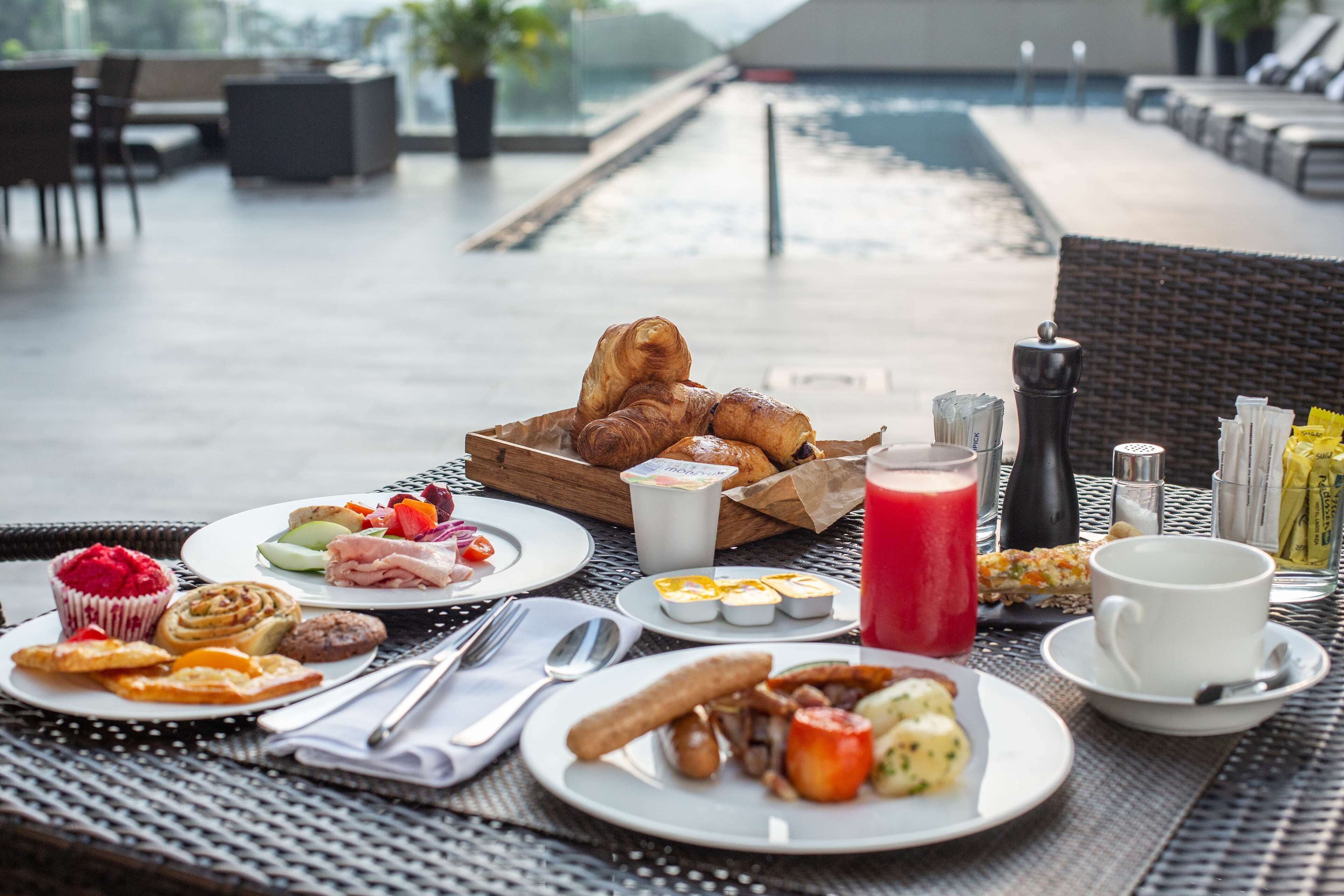 A breakfast buffet with croissants, pastries, and various fruits and cheeses on a table near a poolside area.