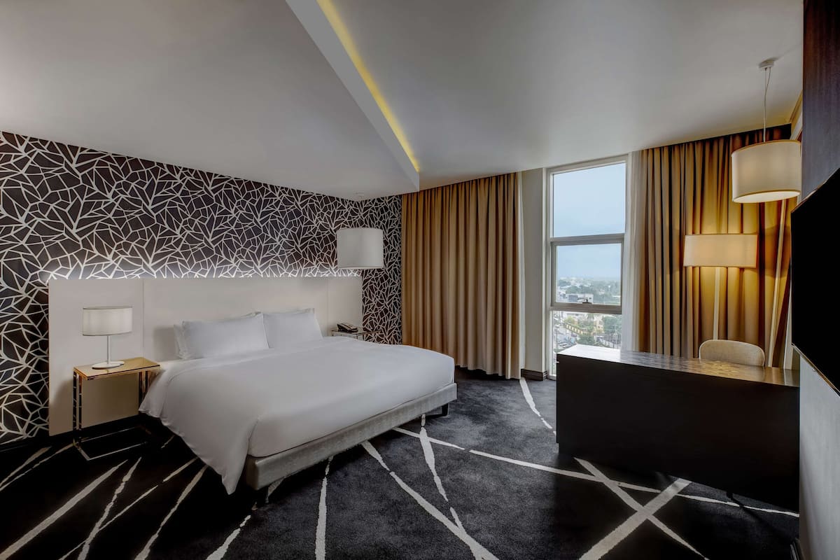A hotel room with a large bed, a desk, a chair, a TV, and a window with curtains.