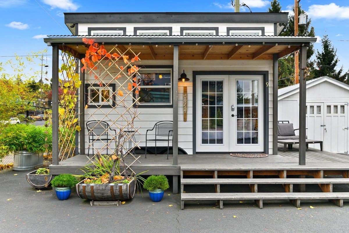 A small, single-story building with a covered porch, a dining area with chairs, and a small garden with potted plants.