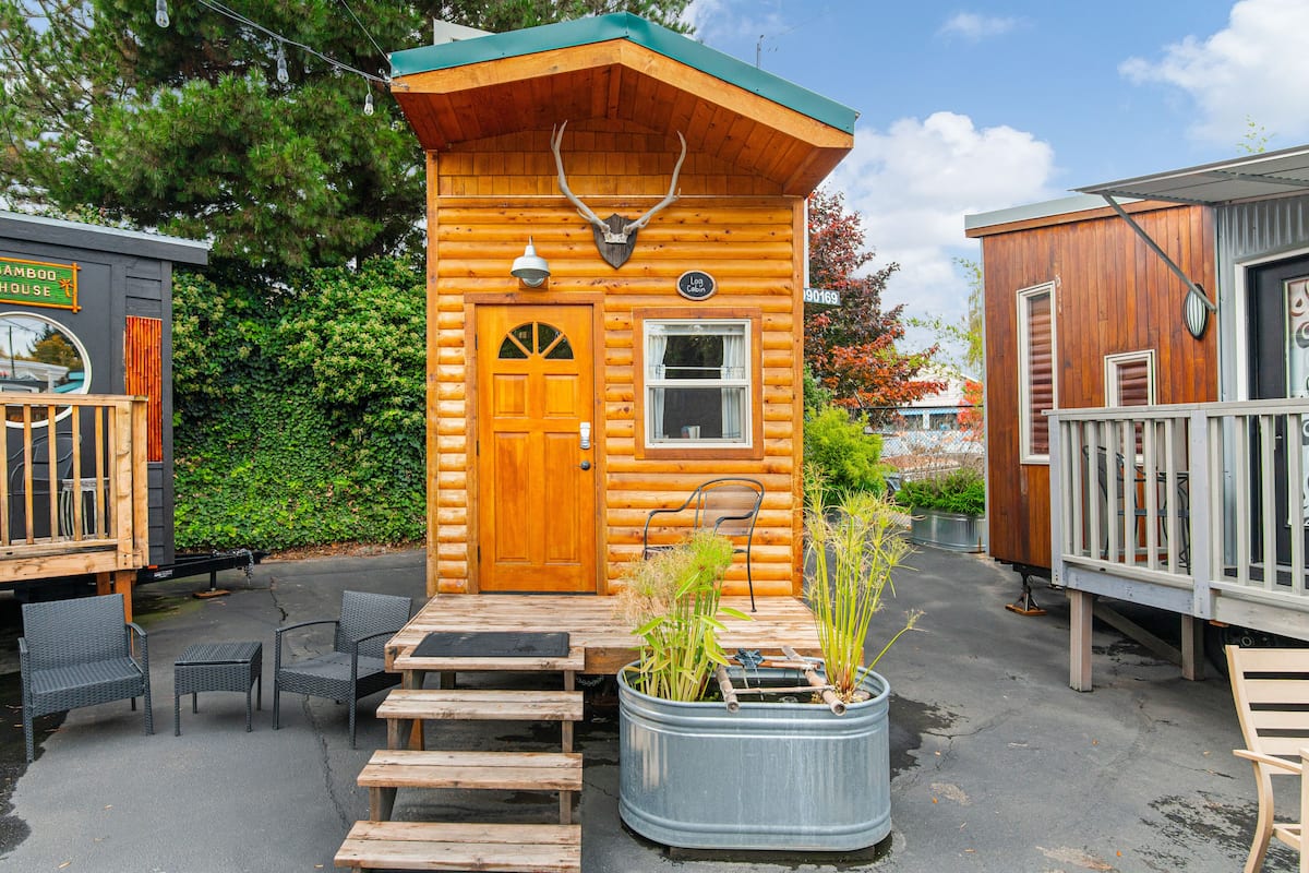 A wooden cabin with a front door, a small window, and a porch with chairs and a table.