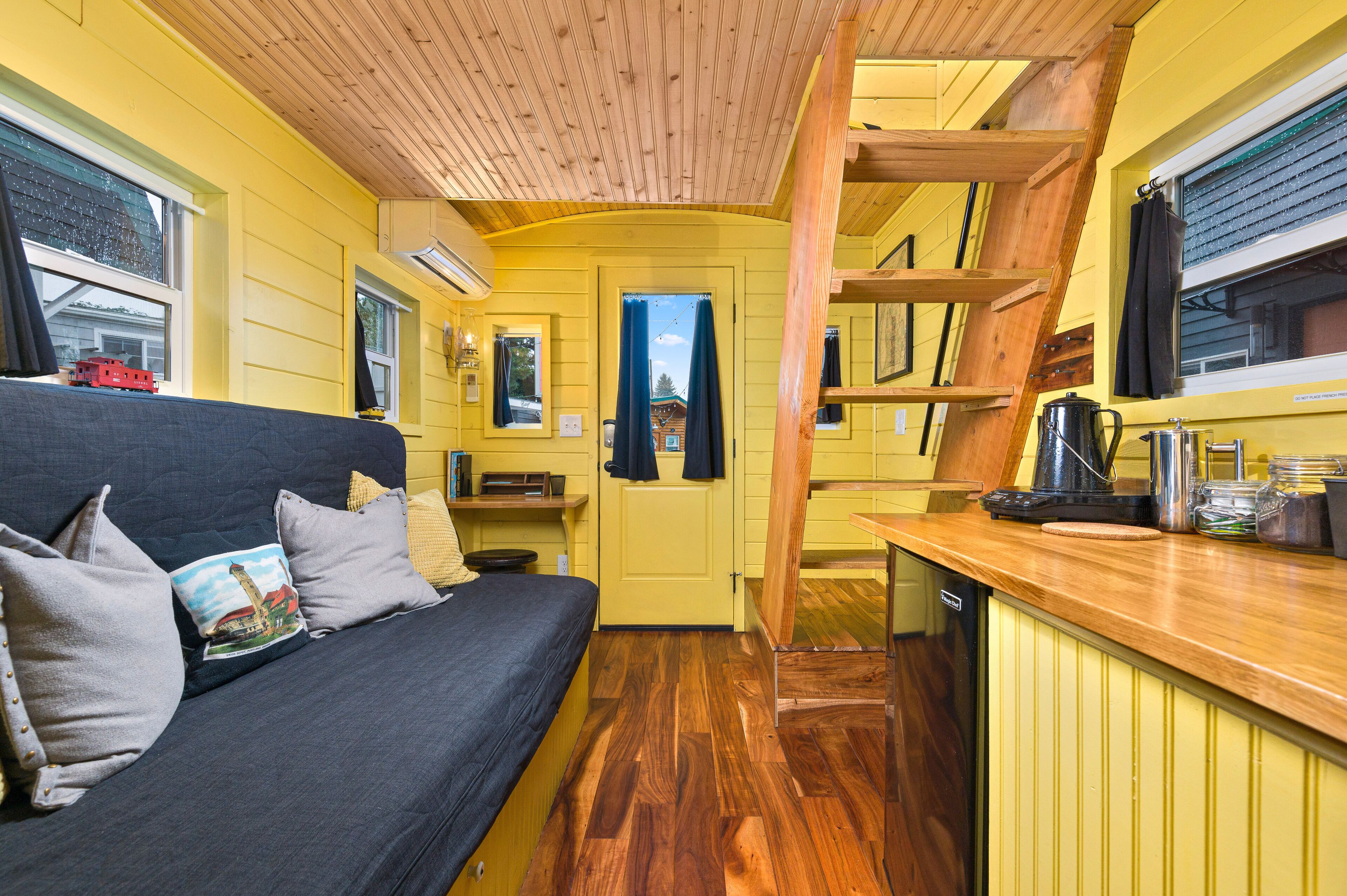Habitación pequeña de color amarillo con cama, kitchenette y vista por una puerta.