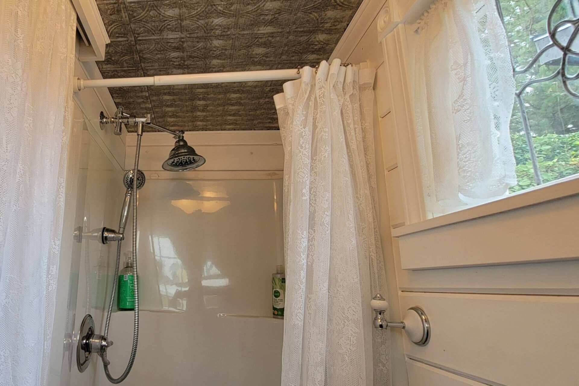 A shower with a handheld showerhead, a glass door, and a window with a view of greenery.