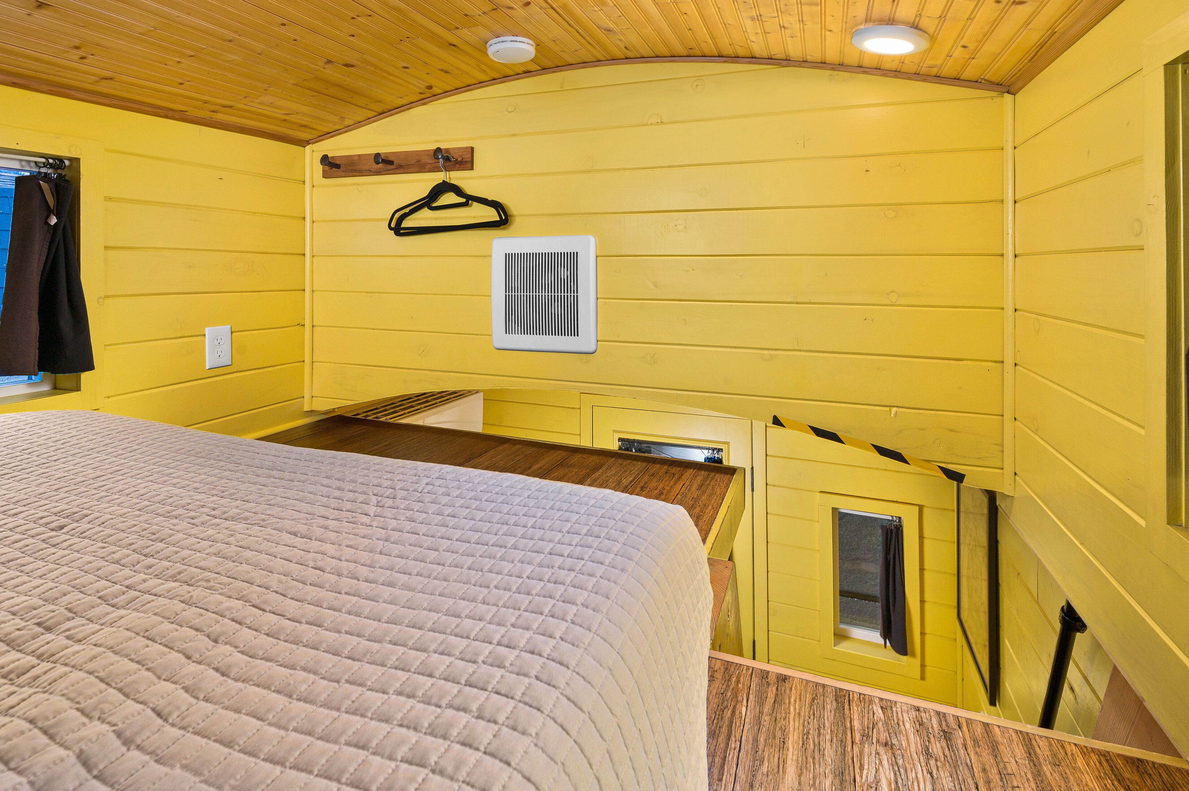 A room with yellow walls, a bed with a quilted cover, a window with a curtain, a wall-mounted heater, and a small window with a curtain.