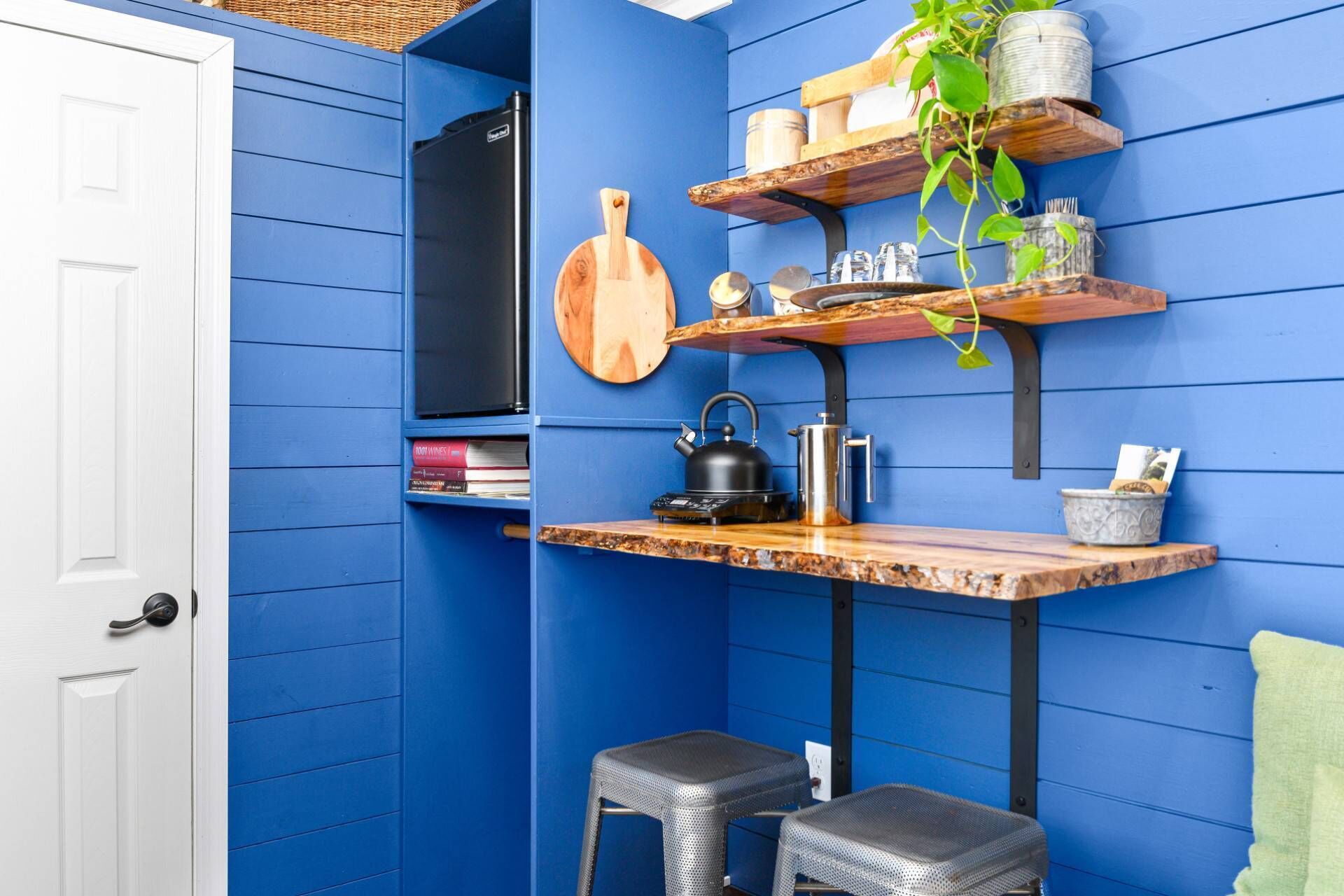 A kitchen with blue walls, a wooden countertop, a kettle, a cutting board, and shelves with kitchen items.