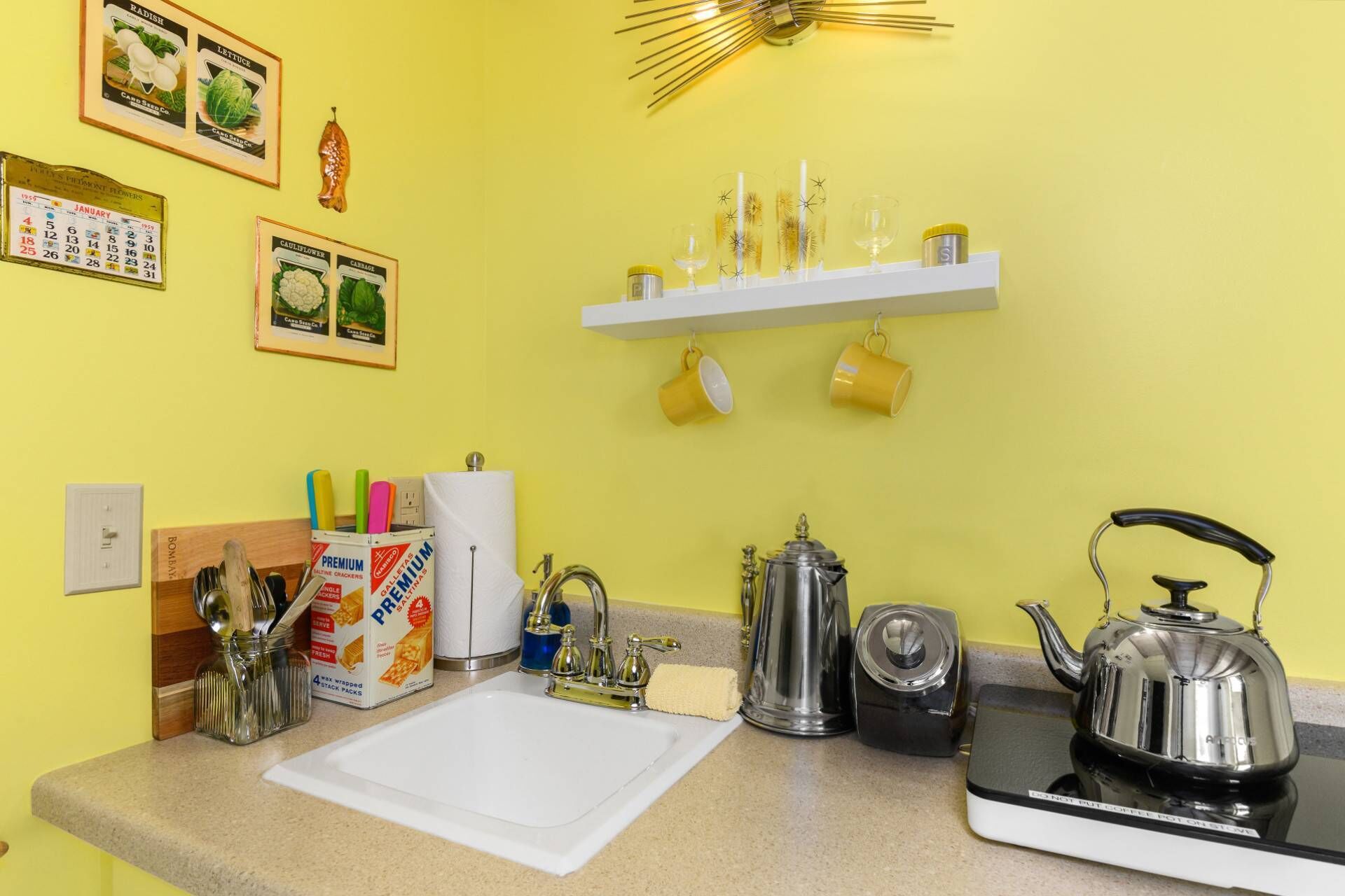 A kitchen with a white countertop, a kettle, a coffee pot, and a shelf with yellow cups.