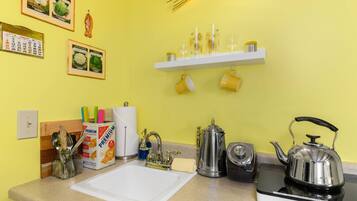 A kitchen with a white countertop, a kettle, a coffee pot, and a shelf with yellow cups.